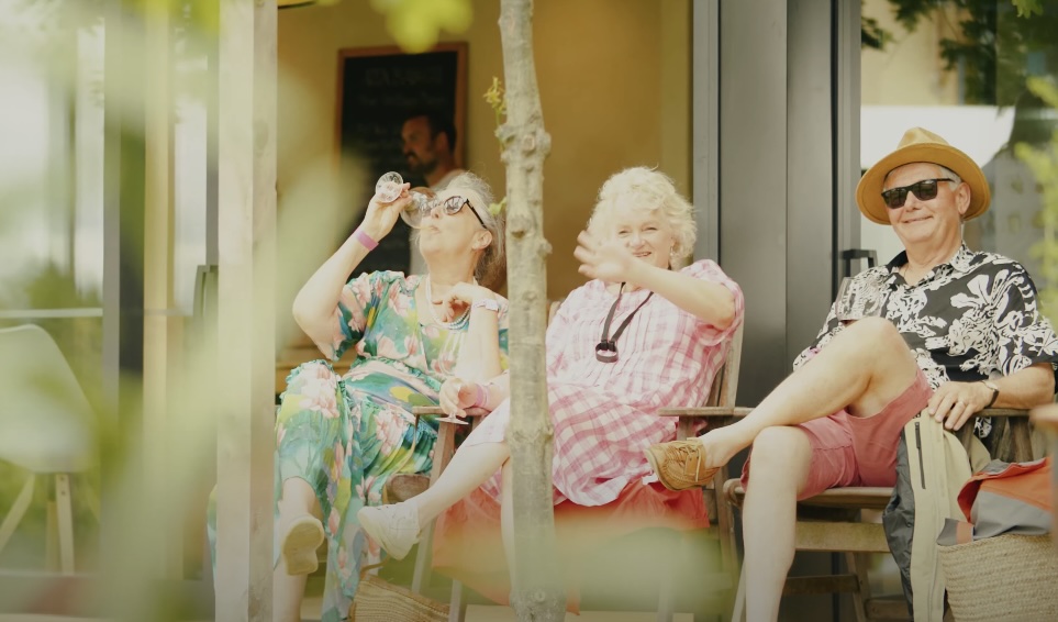 Three elderly individuals sitting, one enjoying a glass of wine